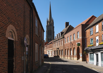 This urban architecture photograph, taken in late morning during spring, features the spire of St James' Church rising above the rooftops in the market town of Louth, Lincolnshire. The scene is characterised by red-brick buildings lining a quiet street, while St James' Church, a prominent landmark in Louth, stands as the focal point in the background. The light conditions suggest a clear spring day, highlighting the historical architecture typical of Lincolnshire towns.